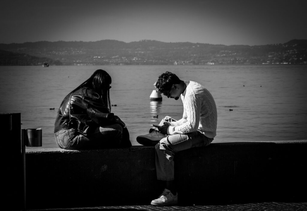 Zwei Menschen sitzen an einem Wasserufer, im Hintergrund Berge und Wasser.